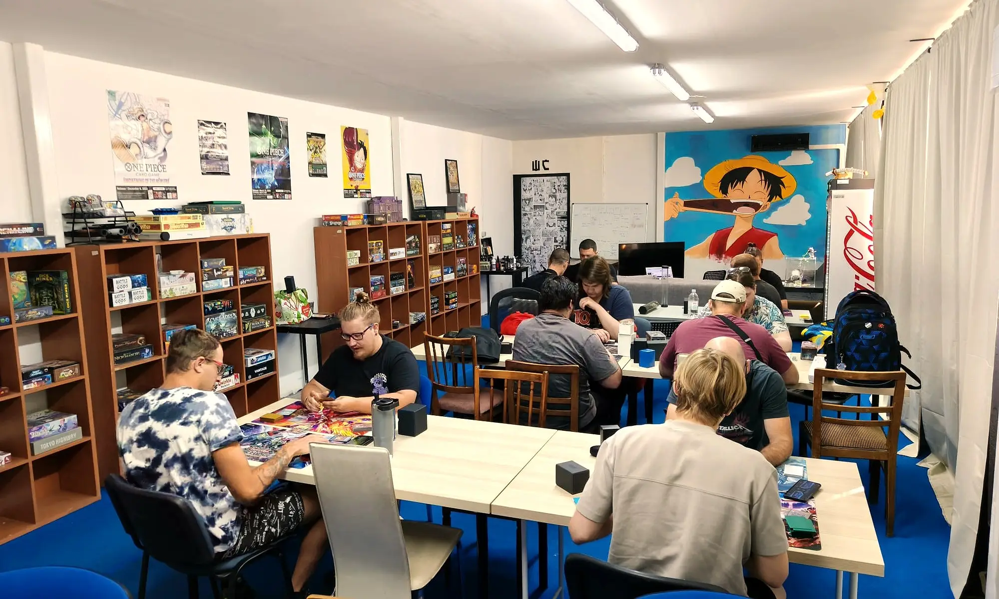 Igrači za stolovima u Card Board Kingdomu, u pozadini police s igrama i mural One Piecea.